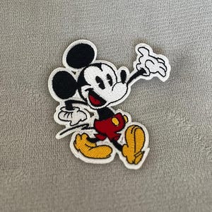 May include: An embroidered fabric patch featuring a classic cartoon mouse character with black ears, a white face, a red mouth, red shorts, and yellow shoes. The character is depicted in a dynamic pose with one arm raised and a white border outline.