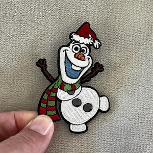 May include: Embroidered patch featuring Olaf the snowman from Frozen, wearing a red Santa hat and a green, red, and white striped scarf. The patch has a white background and black outline.