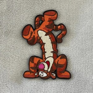 May include: Embroidered patch of Tigger from Winnie the Pooh. The patch depicts Tigger upside down with a happy expression. The patch is orange with brown stripes and a white belly, ideal for decorating clothing or accessories.