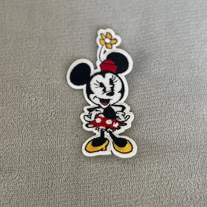 May include: An embroidered patch featuring a cartoon mouse character with black ears, a red hat, and a yellow flower above the head. The character wears a red dress with white polka dots and yellow shoes, outlined in white thread on a fabric base.