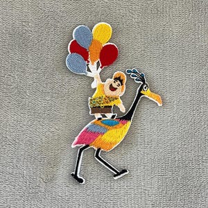 Può includere: Toppa ricamata raffigurante un personaggio del film d'animazione "Up" in sella a un uccello colorato e con dei palloncini. La toppa ha un bordo bianco su sfondo grigio. L'uccello ha un lungo collo e becco.