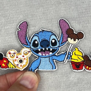 Può includere: Toppa ricamata con Stitch, l'alieno blu di Lilo & Stitch, con un lecca-lecca e dessert. Include una ciambella di Topolino e gelati. La toppa ha un bordo bianco su sfondo grigio.