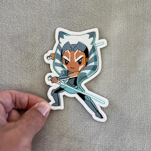 Puede incluir: Parche bordado con un personaje de dibujos animados con piel naranja, colas de cabeza blancas y azules, y dos sables de luz azules. El personaje lleva un atuendo verde azulado oscuro y está en posición de combate. El parche tiene un borde blanco.