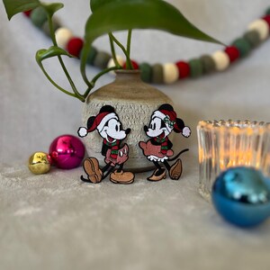 以下が含まれることがあります： ミッキーマウスとミニーマウスをフィーチャーしたクリスマスをテーマにした刺繍パッチのペア。ミッキーは赤と緑のサンタ帽とスカーフを着用し、ミニーは赤と緑のリボンとスカーフを着用しています。パッチは、鉢植えの植物とオーナメントを背景にした白い表面にあります。