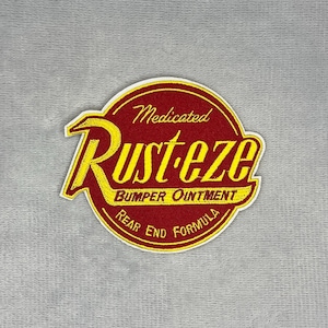 Puede incluir: Un parche bordado circular con fondo rojo y borde amarillo. El texto dice Medicated Rust-eze BUMPER OINTMENT REAR END FORMULA en letras amarillas. El diseño está centrado y tiene un contorno amarillo distintivo.