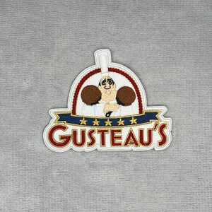 Może przedstawiać: Haftowana naszywka z logo restauracji Gusteau's. Wzór przedstawia postać kucharza w białej czapce, niebieski baner z pięcioma złotymi gwiazdkami oraz czerwony napis GUSTEAU'S, całość zamknięta w białej i czerwonej obwódce w kształcie łuku.