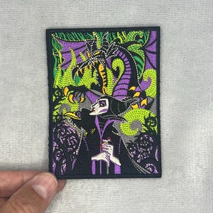 Op de afbeelding: Een rechthoekige geborduurde patch met een figuur in een zwart en paars gewaad met een hoge kraag, staand naast een zwarte vogel. Daarachter bevindt zich een grote paarse en groene draak met ingewikkelde schubben en groene vlammen.