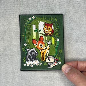May include: A rectangular embroidered patch featuring a forest scene with a fawn, a skunk, a rabbit, and an owl. The design includes green foliage, white flowers, and a butterfly, all rendered in vibrant thread colors on a dark green background.