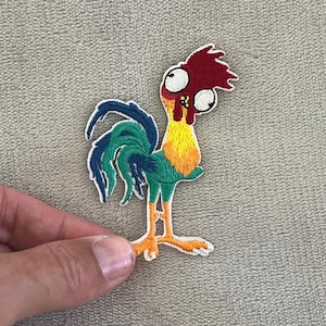 Può includere: Una toppa ricamata colorata raffigurante un gallo dei cartoni animati con cresta rossa, collo giallo e piume della coda verdi e blu. Il gallo ha grandi occhi e zampe arancioni. La toppa è tenuta da una mano.