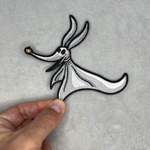 Könnte beinhalten: Gestickter Aufnäher mit Zero, dem Geisterhund aus The Nightmare Before Christmas. Der Aufnäher ist hauptsächlich weiß und grau mit schwarzen Konturen, einem rosa Kragen und einer goldenen Nase. Der Aufnäher wird von einer Hand gehalten.