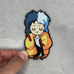 Peut inclure: Écusson brodé représentant un personnage de dessin animé aux cheveux bleus et blancs, portant une veste orange et une robe noire et blanche. L'écusson a une bordure noire et est tenu par une main.