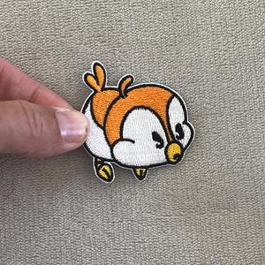 May include: Embroidered patch featuring a cartoon penguin with orange, white, and yellow accents. The penguin has a black outline and is set against a neutral background. The patch is likely for clothing or accessories.