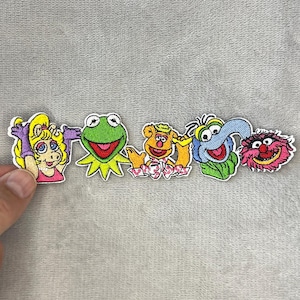 Puede incluir: Una fila de parches bordados con personajes de los Muppets. Los parches incluyen a Miss Piggy, Kermit la Rana, Fozzie Bear, Gonzo y un monstruo rojo. Cada parche tiene un borde blanco.