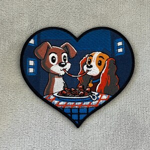 May include: Embroidered patch in the shape of a heart featuring two cartoon dogs sharing spaghetti. The dogs are brown and orange, set against a blue background with white window details. The patch has a black border.