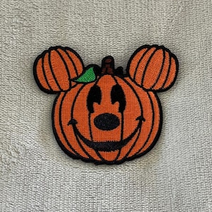 May include: Embroidered patch featuring a smiling pumpkin with Mickey Mouse ears. The pumpkin is orange with black details for the eyes, nose, and mouth. The ears are also orange with black outlines, and a small green leaf is visible.