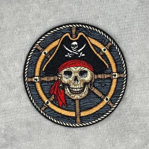 May include: Embroidered patch featuring a pirate skull wearing a red bandana and a black hat with a skull and crossbones. The skull is centered within a ship's wheel design, with a navy blue and gold color scheme.