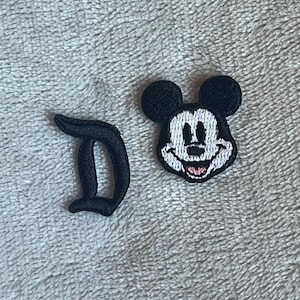 May include: Two embroidered patches: a black letter "D" and a Mickey Mouse head. The Mickey Mouse patch features a white face, black ears, and a pink tongue. These patches are likely for clothing or accessories.