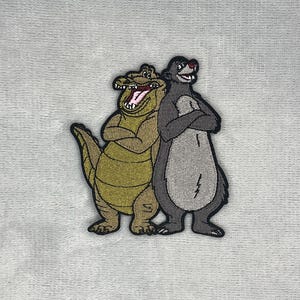 May include: An embroidered patch featuring a cartoon alligator with olive green skin and an open mouth standing next to a grey bear with a light grey belly. Both characters have their arms crossed and are outlined in black thread on a light fabric.