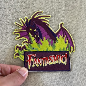 May include: Embroidered patch featuring a purple dragon with green flames and the word "Fantasmic!" in red and yellow. The patch has a dark purple border and is likely for clothing or accessories.