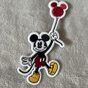 Puede incluir: Un parche bordado para planchar con Mickey Mouse sosteniendo un globo rojo con orejas de Mickey Mouse. El parche es blanco con contornos negros y presenta a Mickey Mouse con sus clásicos pantalones cortos rojos y zapatos amarillos.