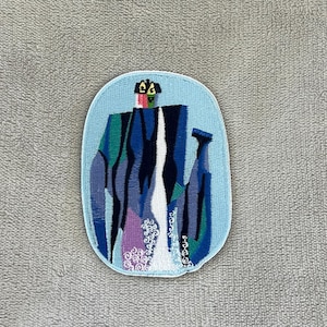 May include: An embroidered patch with a light blue background and a mountain landscape design. The patch features shades of blue, green, purple, and white, with a small house at the top. The patch has a white border.