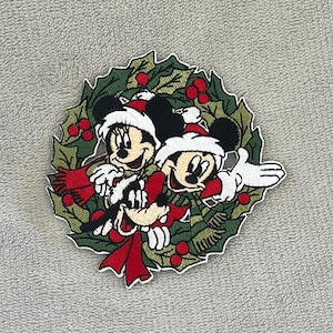 May include: Embroidered patch featuring Mickey Mouse, Minnie Mouse, and Goofy in a Christmas wreath. The characters wear Santa hats and red scarves. The wreath is green with red berries, perfect for holiday decor.