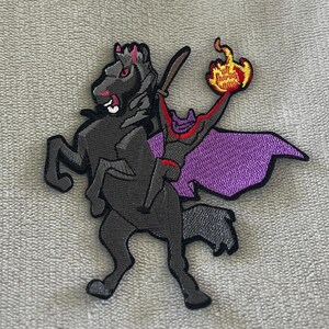 May include: Embroidered patch featuring the Headless Horseman riding a black horse. The horse and rider are in a dynamic pose, with the rider holding a flaming torch. The patch has purple, red, orange, and black colors.