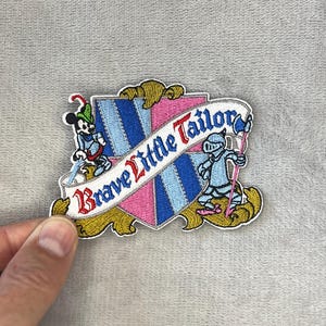 May include: Embroidered patch featuring Mickey Mouse and a knight, with the text "Brave Little Tailor" on a shield design. The patch has a pink and blue color scheme with gold accents.