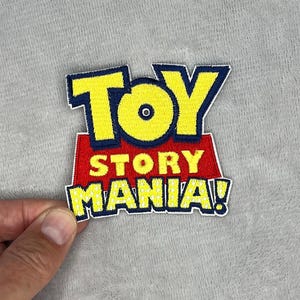 Puede incluir: Parche bordado con el texto "TOY STORY MANIA!" en amarillo, rojo y azul. El parche tiene un borde blanco y está sobre un fondo gris. El parche está sujeto por una mano.