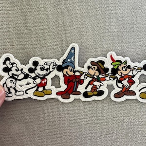 May include: A white-bordered embroidered patch featuring six distinct Mickey Mouse figures in a horizontal row. The characters are depicted in various outfits, including a sorcerer robe with a blue hat, a brown explorer hat, and a green feathered cap.