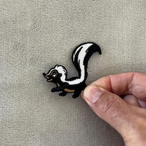 Könnte beinhalten: Gestickter Aufnäher mit einem Cartoon-Stinktier. Das Stinktier ist schwarz mit einem weißen Streifen auf dem Rücken und einem weißen Streifen auf dem Kopf. Der Aufnäher hat eine goldfarbene Umrandung und wird von einer Hand gehalten.