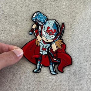 May include: Embroidered patch featuring a cartoon version of Thor, holding his hammer. The patch has a red cape, blue and silver armor, and a helmet with a red design. The patch is on a gray background.