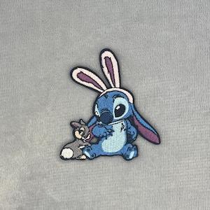 May include: An embroidered patch featuring a blue alien character wearing pink and white rabbit ears, sitting next to a small grey and white rabbit. The patch has a black border and detailed stitching in shades of blue, pink, grey, and white.