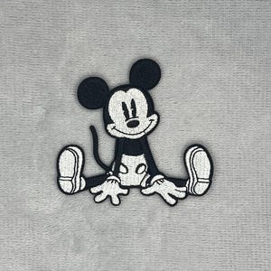 以下が含まれることがあります： 足を前に伸ばして座っている漫画のネズミのキャラクターが描かれた、黒と白の刺繍ワッペン。キャラクターには大きな丸い耳、笑顔の顔、地面に置かれた手袋をした手があり、詳細な糸のステッチで表現されています。