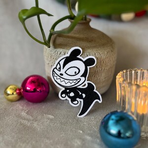 Puede incluir: Parche bordado en blanco y negro de un monstruo de dibujos animados con una sonrisa de dientes. El monstruo lleva una camisa a rayas blancas y negras y tiene una cola negra.