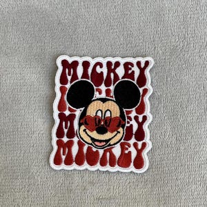 Pode incluir: Patch bordado com Mickey Mouse usando óculos de sol vermelhos em forma de coração. O patch tem as palavras "Mickey" em um padrão repetido na cor bordô, com uma borda branca.