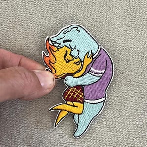 May include: Embroidered patch featuring two cartoon figures embracing. One figure is blue with a purple shirt, the other is yellow with flames for hair and a red plaid skirt. The patch has a white border.