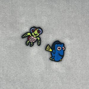 Könnte beinhalten: Zwei gestickte Aufnäher mit Zeichentrickfiguren. Ein Aufnäher zeigt eine kleine grüne Meeresschildkröte mit rosa Panzer und großen Augen. Der zweite Aufnäher zeigt einen blauen Fisch mit gelben Flossen und großen Augen.