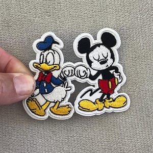 May include: Embroidered patch featuring Donald Duck and Mickey Mouse bumping fists. Donald Duck is wearing a blue sailor suit and hat, while Mickey Mouse is in red shorts. Both characters have yellow shoes and are outlined in white.