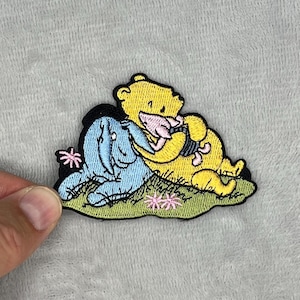 May include: Embroidered patch featuring Winnie the Pooh, Piglet, and Eeyore. The patch shows the characters in a playful embrace, set against a green grassy area with pink flowers. The patch is yellow, blue, pink, and black.