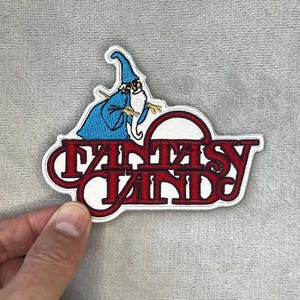 Può includere: Toppa ricamata con un mago in abito blu con una lunga barba bianca e un bastone. La toppa presenta la scritta "FANTASY LAND" in lettere rosse con bordo bianco. Sfondo grigio.