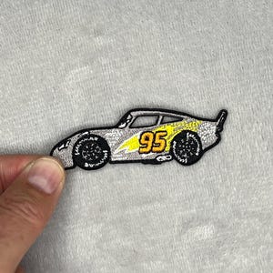 May include: A silver and yellow embroidered patch in the shape of a race car. The number 95 is displayed in yellow on the side of the car. The car has black wheels and a black outline.