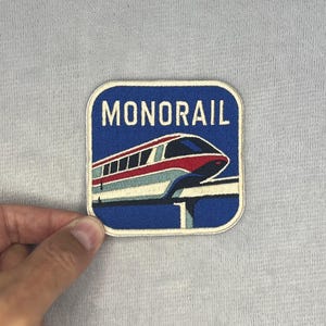 May include: A square embroidered patch with rounded corners featuring a blue background and the word MONORAIL in white capital letters. The design depicts a white, red, and blue monorail train traveling on a track, with the number 1 visible below.