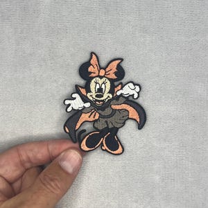 May include: An embroidered patch featuring a cartoon mouse character dressed as a vampire. The character wears a black and orange cape, a matching bow, and a dark dress. The design includes white gloves and black shoes, with a black outline throughout.