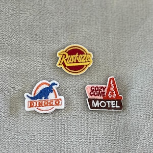May include: Three embroidered patches featuring designs from a popular animated film. The patches include the "Rust-eze" logo, a dinosaur with "Dinoco" text, and the "Cozy Cone Motel" sign.