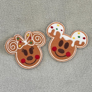 以下が含まれることがあります： ミッキーマウスの頭の形をした、ジンジャーブレッド風のパッチが2つ。1つはリボン、もう1つは白いアイシングが付いています。どちらも茶色の顔、赤い頬、白い輪郭です。クリスマスのクラフトに最適です。