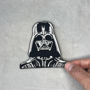 May include: Black and white embroidered patch featuring the helmet of Darth Vader. The patch is a detailed depiction of the iconic Star Wars character's helmet, with intricate stitching and a bold design.