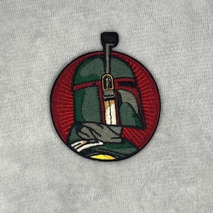 May include: Embroidered patch depicting a science fiction character's helmet. The circular patch has a red border and a detailed image of the helmet in green, grey, and white. The patch is designed to be ironed onto fabric.