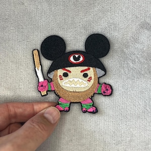 Pode incluir: Patch bordado com um personagem de desenho animado com chapéu e orelhas pretas, corpo marrom e detalhes rosa e verde. O personagem segura um objeto dourado e branco. O patch tem uma boca branca e irregular e um design de olho vermelho.