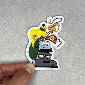 Könnte beinhalten: Gestickter Aufnäher mit Cartoon-Figuren, die übereinander gestapelt sind. Der Aufnäher zeigt eine gelbhaarige Figur in einem grünen Outfit, einen blauen Toaster mit einem lächelnden Gesicht und einen grauen Staubsauger.